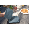 Hand hält graublauen rechteckigen Behälter mit Deckel mit Noppen auf einem Holztisch; daneben Teller mit Croissant, Marmeladenglas und eine kleine Sukkulente