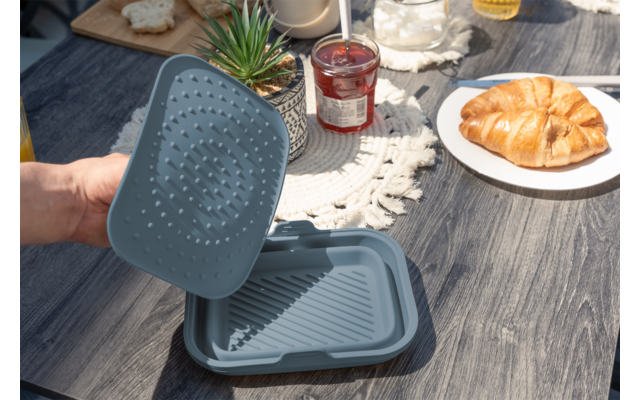 Hand hält graublauen rechteckigen Behälter mit Deckel mit Noppen auf einem Holztisch; daneben Teller mit Croissant, Marmeladenglas und eine kleine Sukkulente