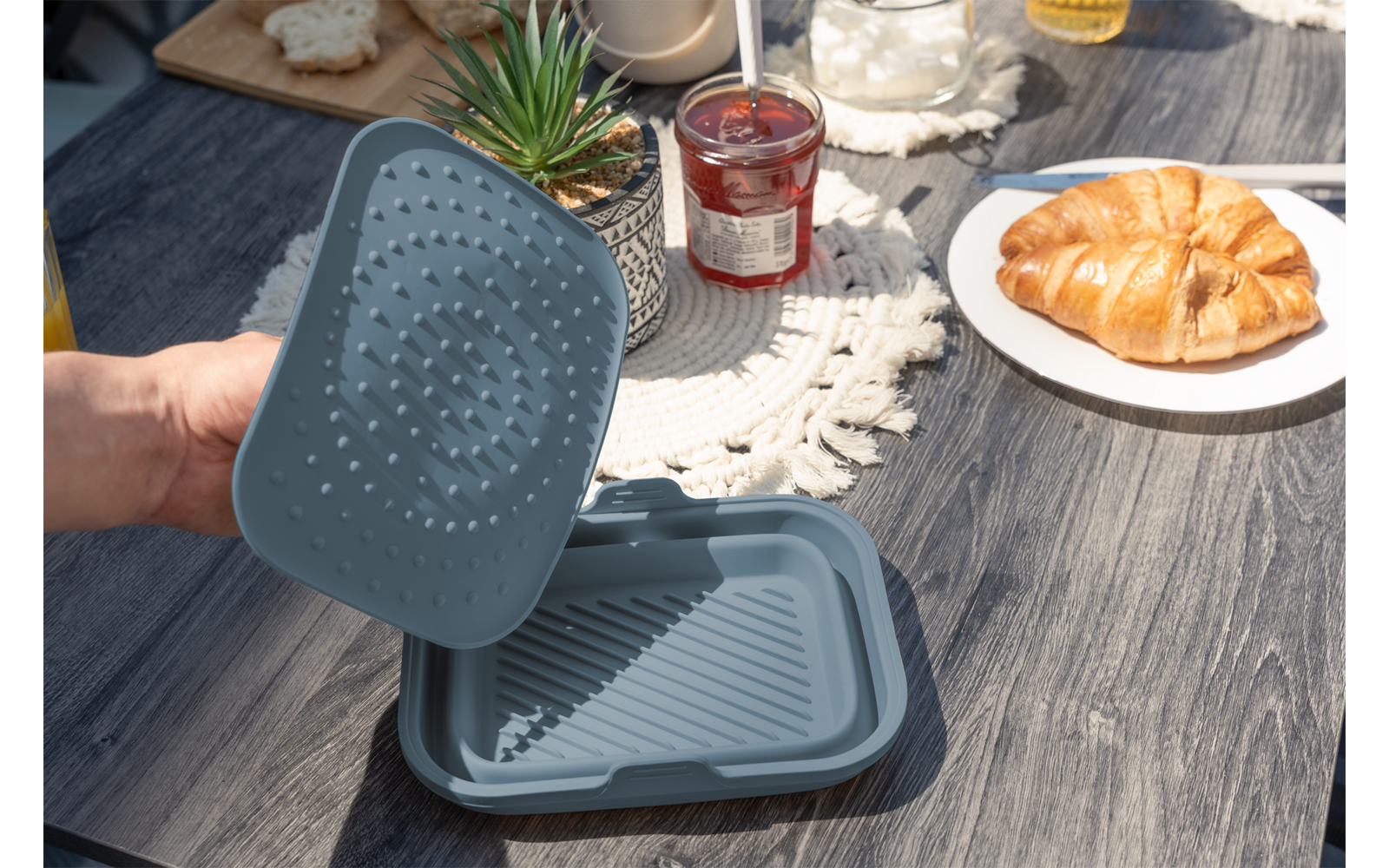 Hand hält graublauen rechteckigen Behälter mit Deckel mit Noppen auf einem Holztisch; daneben Teller mit Croissant, Marmeladenglas und eine kleine Sukkulente