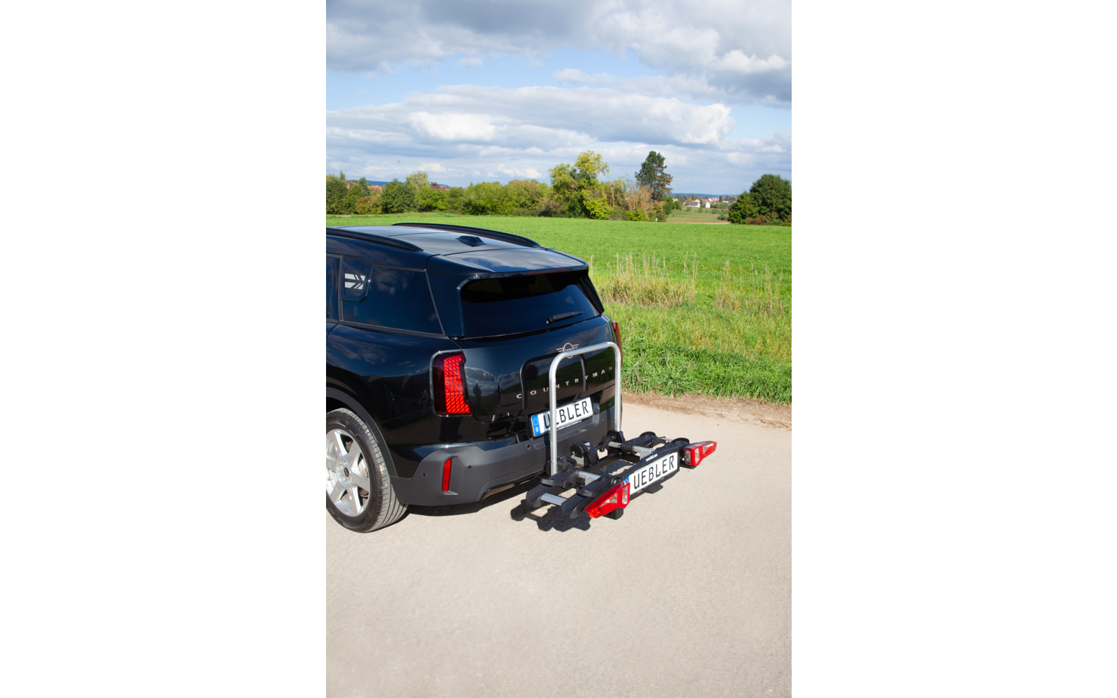 Heck eines dunklen SUVs mit angebrachtem Fahrradträger, im Hintergrund Wiese, Bäume und bewölkter Himmel