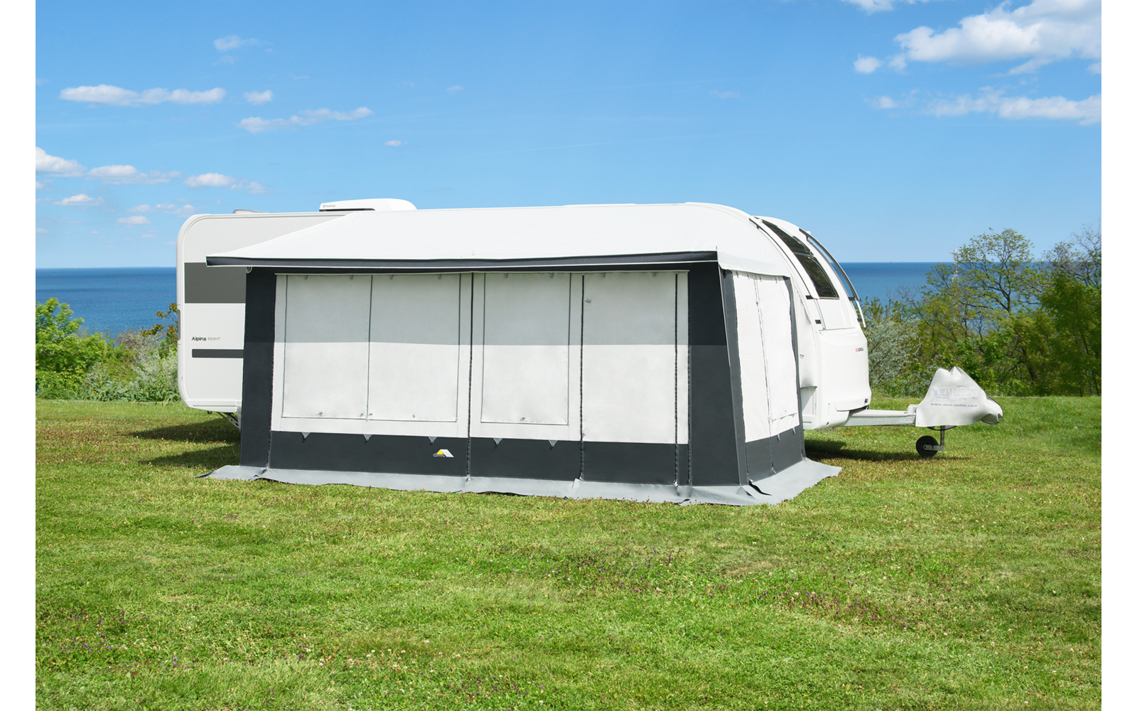 Weißer Wohnwagen mit grauem Vorzelt auf einer Wiese, dahinter das Meer, blauer Himmel mit vereinzelten Wolken und einige Büsche/kleine Bäume