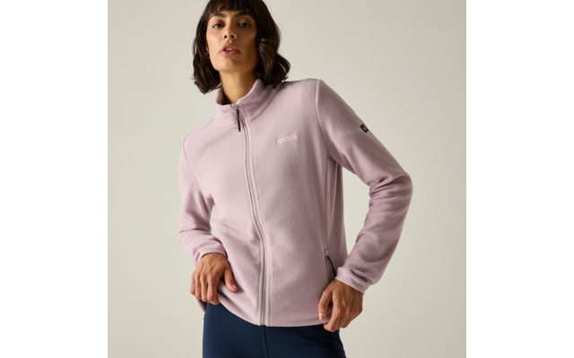 Persona con giacca in pile rosa chiaro con cerniera e pantaloni scuri, in piedi davanti a uno sfondo chiaro uniforme, mani sui fianchi