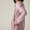Vista laterale di una persona che indossa una giacca in pile rosa chiaro, mano nella tasca con zip, mento e bocca visibili, piccolo marchio nero sulla manica, sfondo chiaro