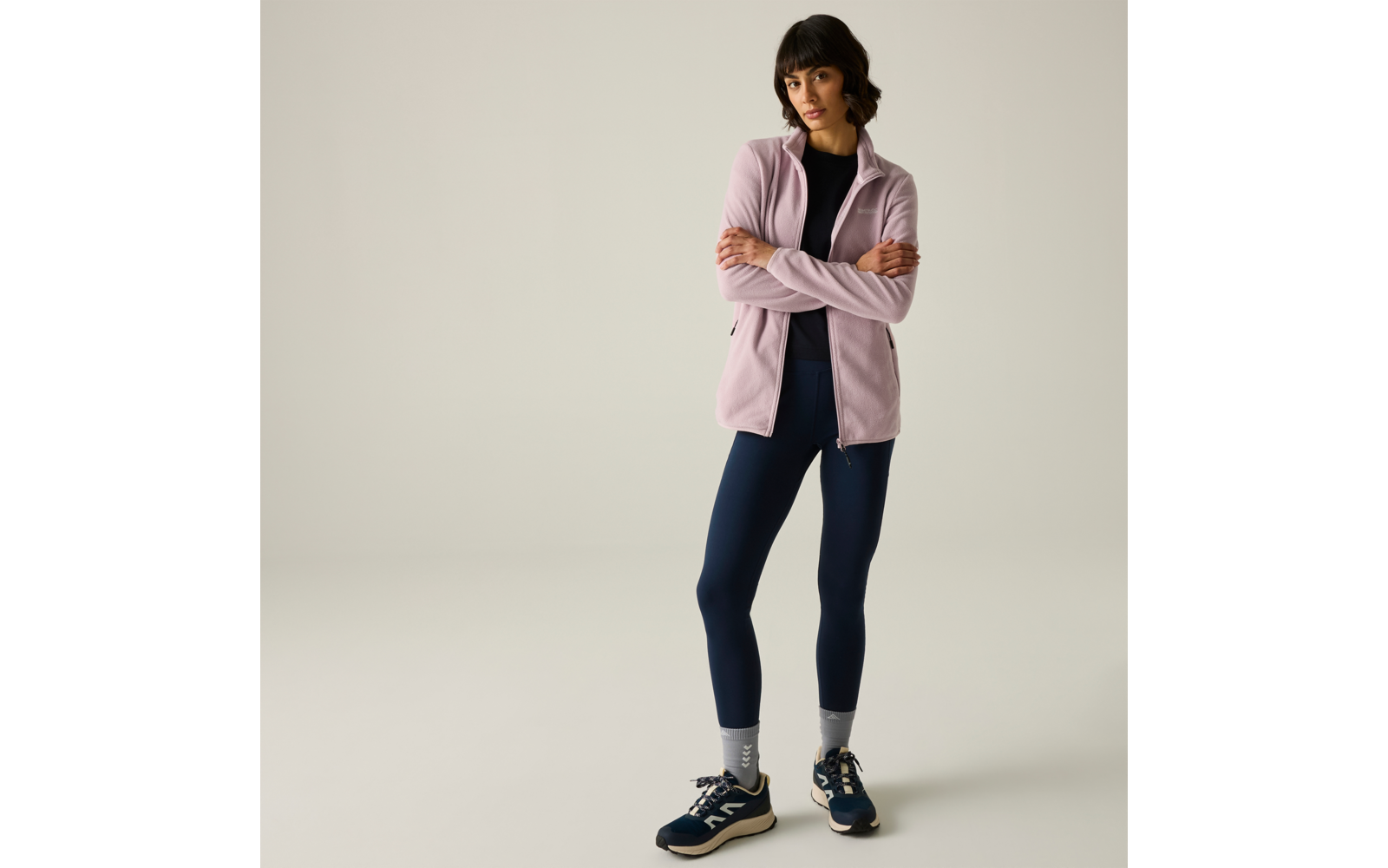 Persona in piedi con le braccia incrociate, indossa una giacca lilla chiaro con zip, top nero, leggings blu scuro, calzini grigi e sneakers blu scuro, sfondo chiaro uniforme