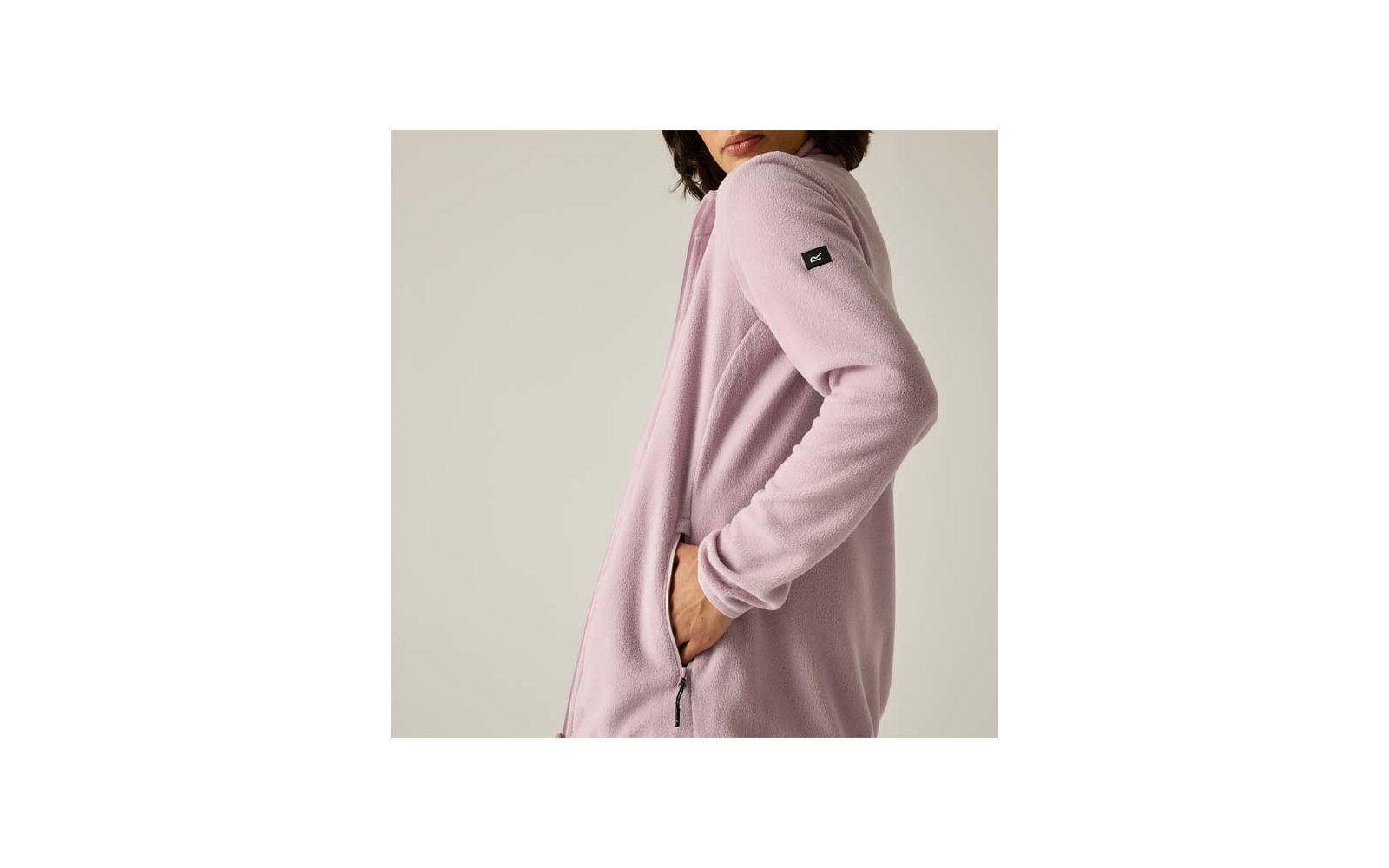 Vista laterale di una persona che indossa una giacca in pile rosa chiaro, mano nella tasca con zip, mento e bocca visibili, piccolo marchio nero sulla manica, sfondo chiaro
