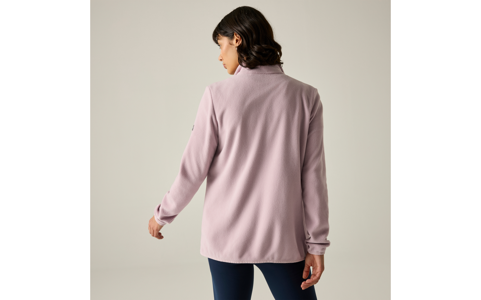 Persona vista da dietro con capelli corti e scuri, indossa una giacca rosa chiaro e pantaloni scuri aderenti su sfondo chiaro neutro
