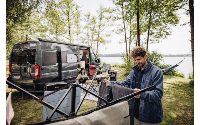 Un homme étend du linge, avec un camping-car et des arbres près d'un lac en arrière-plan