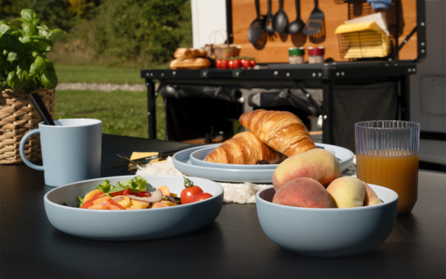 Table extérieure noire avec deux bols bleu clair (un avec salade, un avec pêches), assiette de croissants, verre de jus d'orange et tasse bleu clair ; en arrière-plan, cuisine de camping ouverte avec ustensiles suspendus et panier à pain