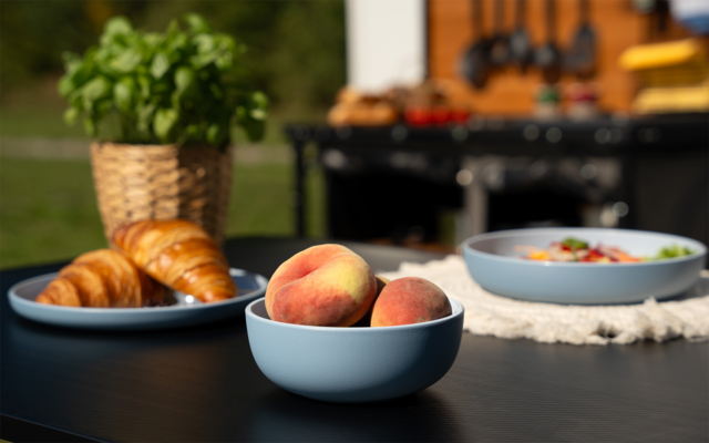 Bol bleu contenant trois pêches sur une table foncée, à côté d'une assiette bleue avec des croissants, un autre bol bleu avec des aliments variés, un pot tressé avec une plante verte et un grill flou avec des ustensiles de cuisine en arrière-plan