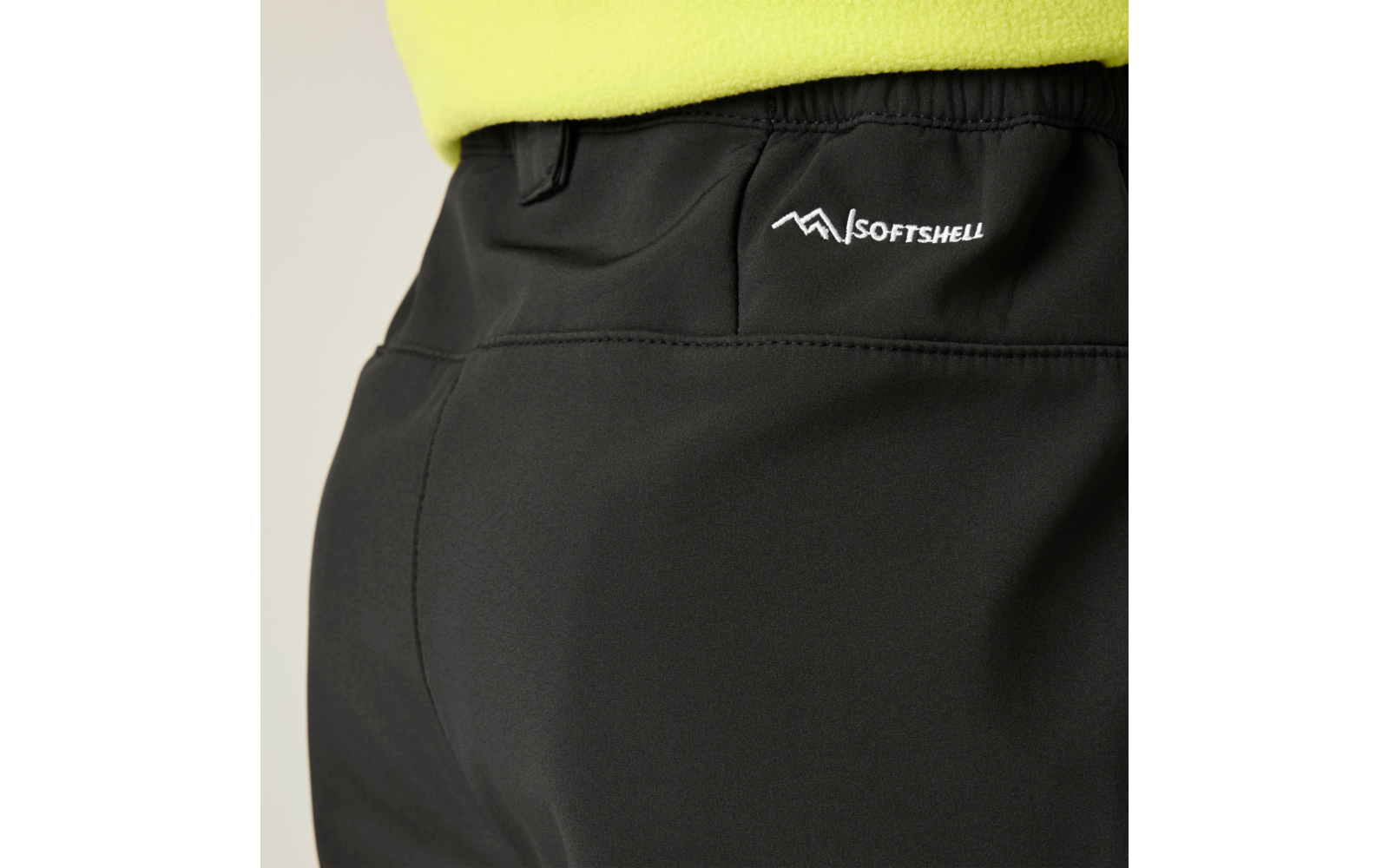 Nahaufnahme der Rückseite einer schwarzen Softshell-Hose mit elastischem Bund, sichtbarer Naht und weißem SOFTSHELL-Logo rechts; unterer Saum eines gelbgrünen Oberteils