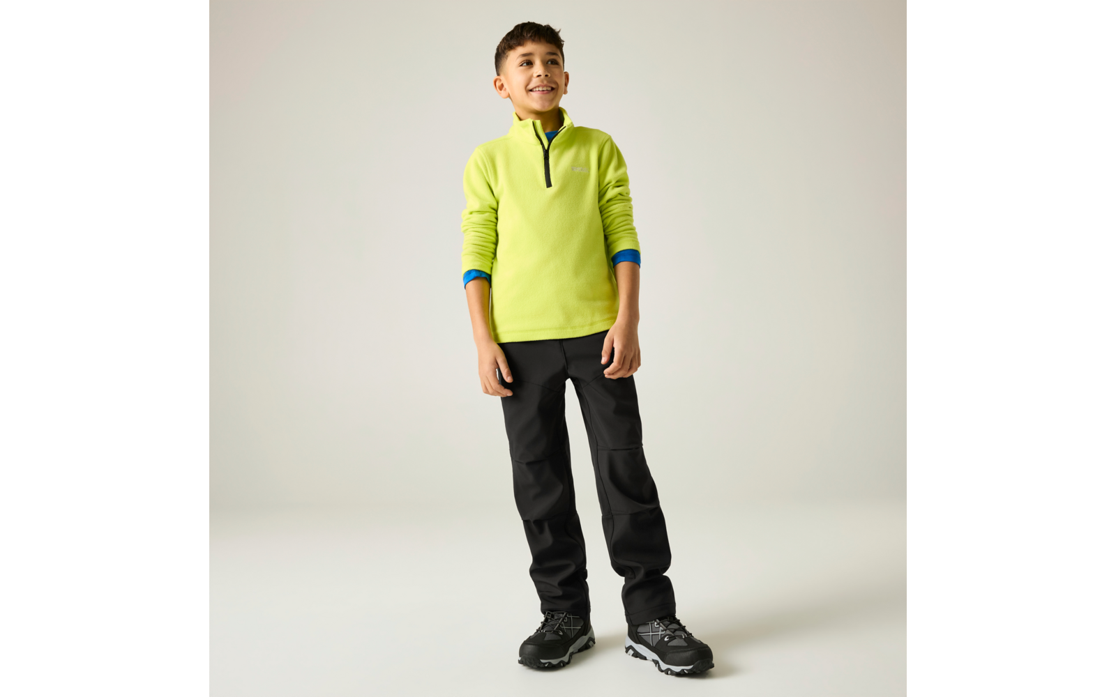 Ganzkörperaufnahme eines lächelnden Jungen, stehend vor hellem Hintergrund, mit neongrünem Pullover mit kurzem Reißverschluss, schwarzer Hose und schwarzen Sportschuhen