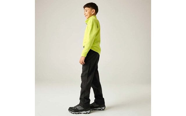Kind seitlich stehend, trägt neongrünes Langarmshirt, schwarze Cargohose und schwarze Sportschuhe vor einfarbig hellem Hintergrund