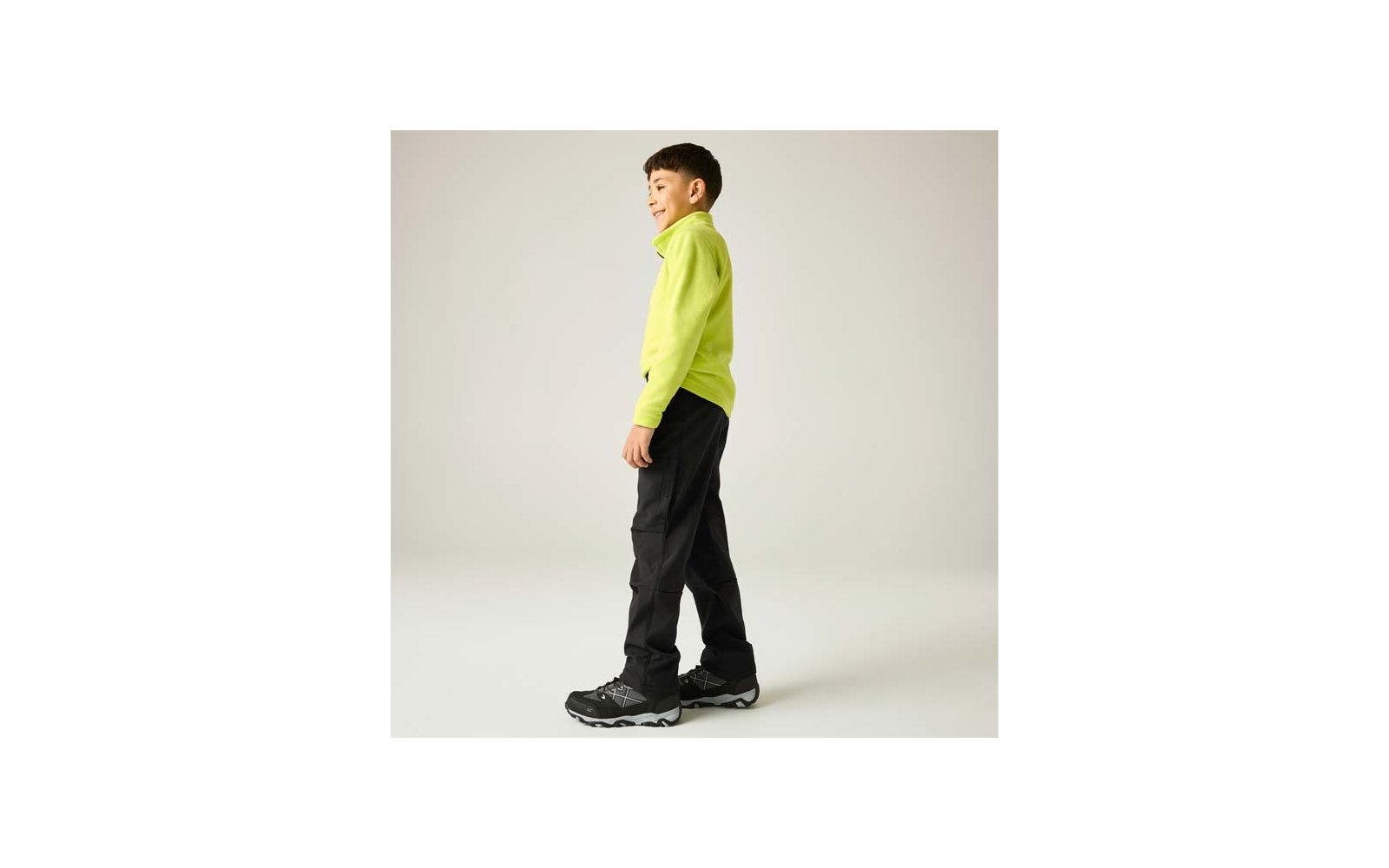 Kind seitlich stehend, trägt neongrünes Langarmshirt, schwarze Cargohose und schwarze Sportschuhe vor einfarbig hellem Hintergrund
