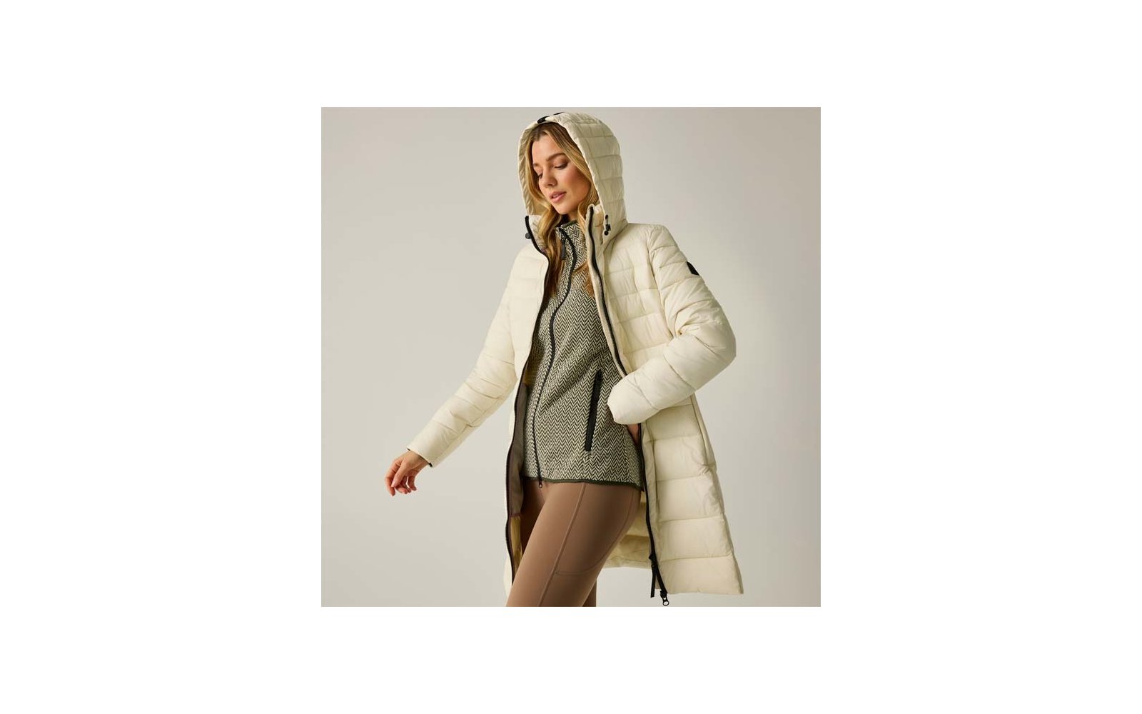 Donna con cappotto trapuntato lungo color crema con cappuccio, aperto sul davanti, maglione grigio strutturato e leggings beige, sfondo neutro
