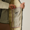 Due mani tengono aperta una giacca imbottita beige, mostrando la fodera interna con piping lilla e un'etichetta bianca; sotto si vede un maglione a motivo bianco e nero e pantaloni marroni