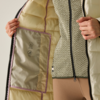 Mani che tengono aperto l'interno di una giacca trapuntata beige chiaro; tasca interna con simboli di cura, cerniera nera, maglia a motivo e pantaloni beige visibili