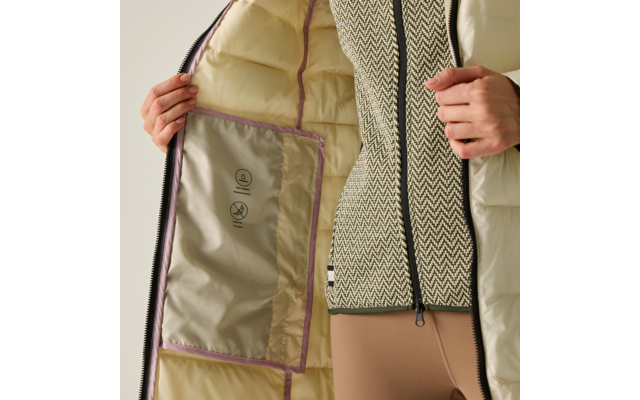 Mani che tengono aperto l'interno di una giacca trapuntata beige chiaro; tasca interna con simboli di cura, cerniera nera, maglia a motivo e pantaloni beige visibili