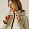 Donna con giacca imbottita beige chiaro con cappuccio che tiene i cordini al collo, cerniera visibile, guarda verso il basso con un leggero sorriso