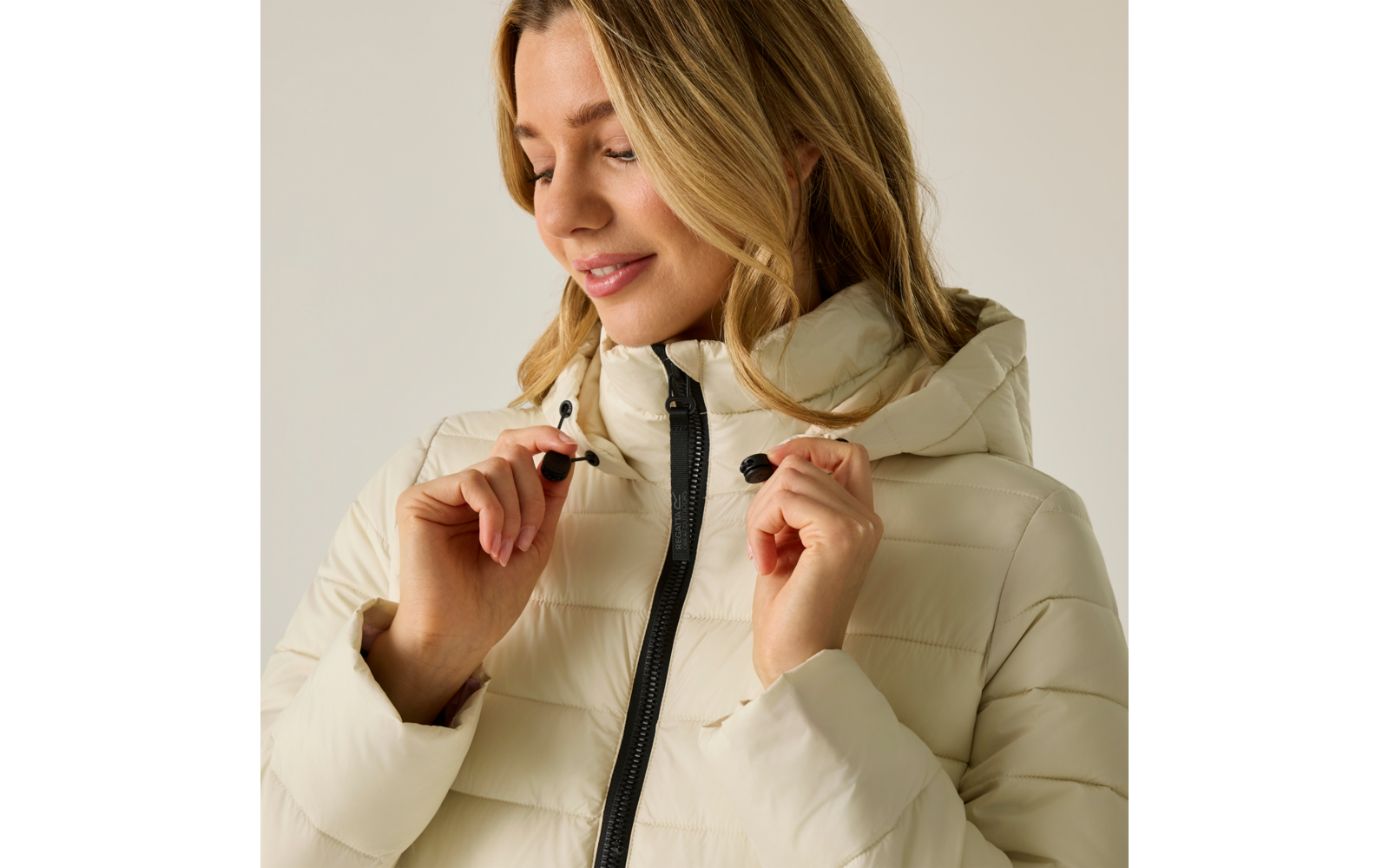 Donna con giacca imbottita beige chiaro con cappuccio che tiene i cordini al collo, cerniera visibile, guarda verso il basso con un leggero sorriso