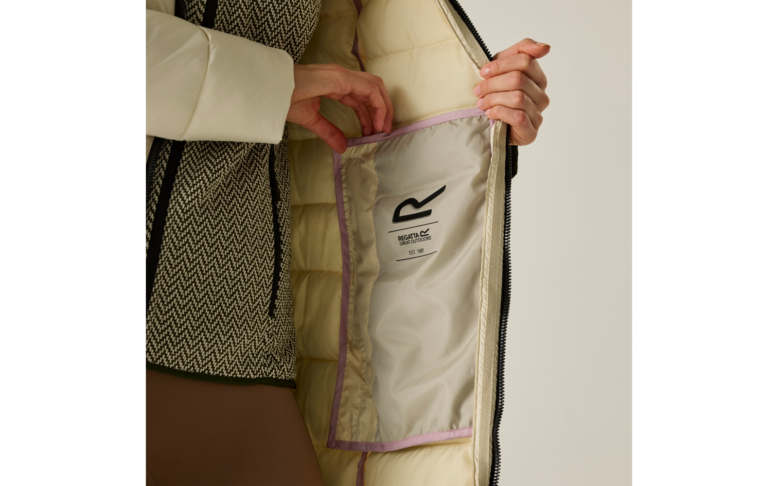 Due mani tengono aperta una giacca imbottita beige, mostrando la fodera interna con piping lilla e un'etichetta bianca; sotto si vede un maglione a motivo bianco e nero e pantaloni marroni