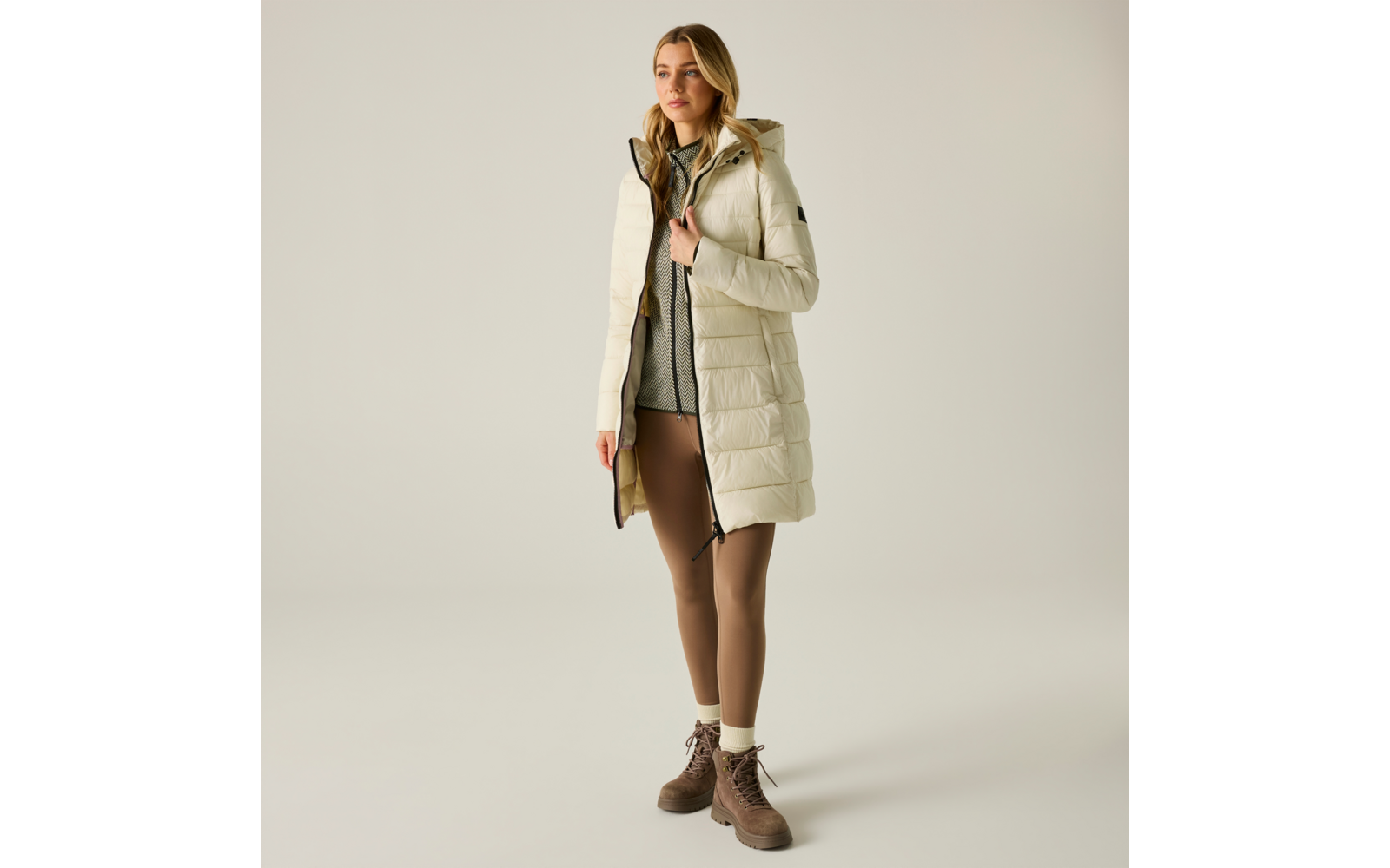 Donna con giacca trapuntata chiara al ginocchio con cappuccio, maglione grigio, leggings marroni e stivaletti marroni stringati su sfondo chiaro
