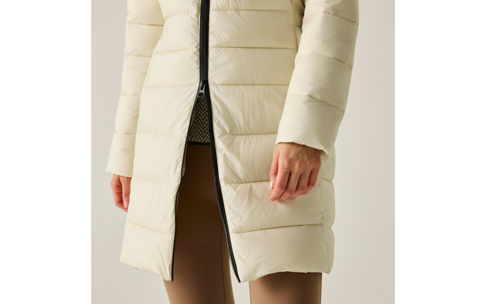 Vista parziale di una persona dalle anche con un cappotto imbottito color avorio e cerniera centrale parzialmente aperta, sotto una maglia con motivo e leggings marroni, mani visibili ai lati, sfondo neutro