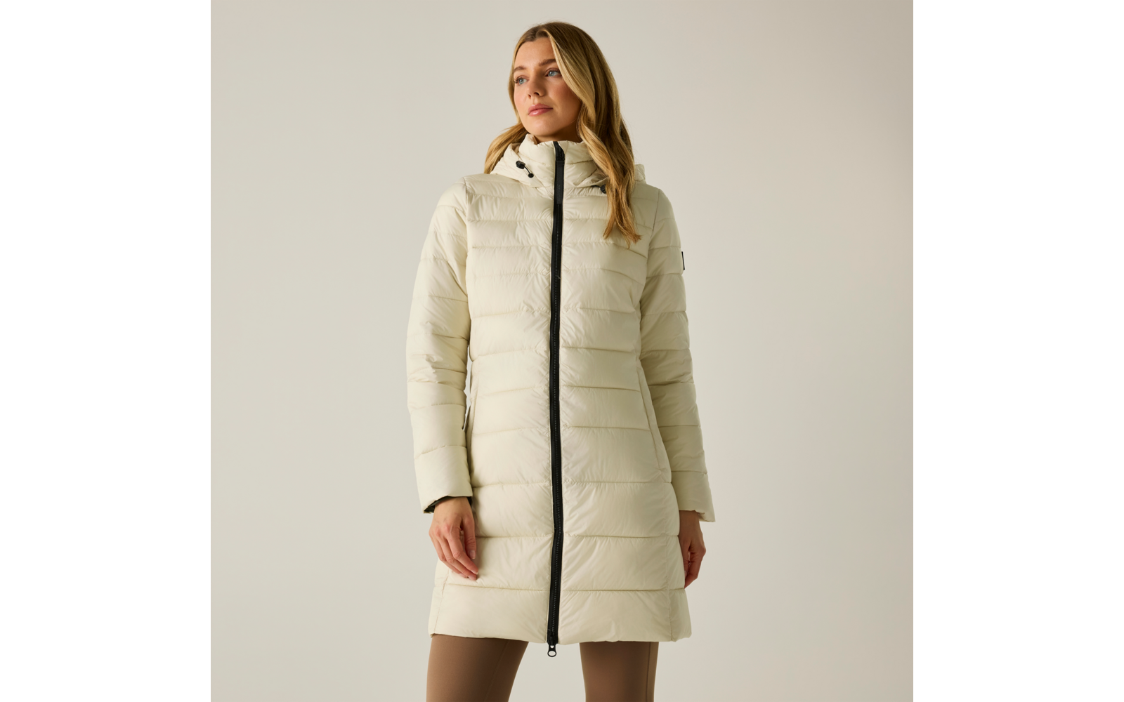 Donna su sfondo chiaro che indossa un cappotto trapuntato beige chiaro con cappuccio e zip nera, lunghezza al ginocchio, pantaloni beige visibili