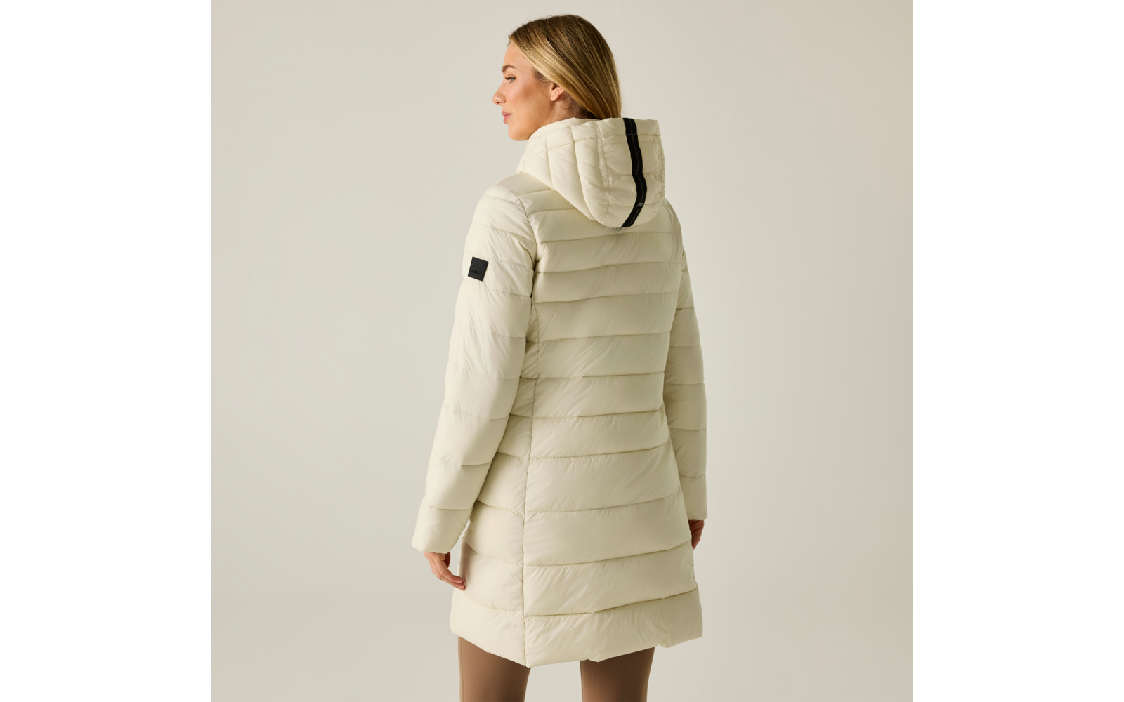 Vista posteriore di una donna con cappotto trapuntato chiaro fino ai fianchi con cappuccio, striscia nera sul retro del cappuccio e toppa nera sulla manica, sfondo chiaro