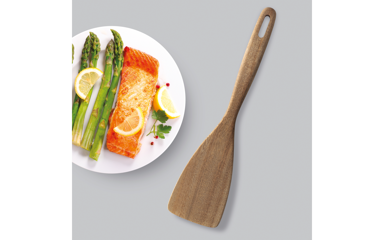 Gebratener Lachs mit Spargel, Zitronenscheiben und Tomaten auf einem weißen Teller neben einem Holzspatel