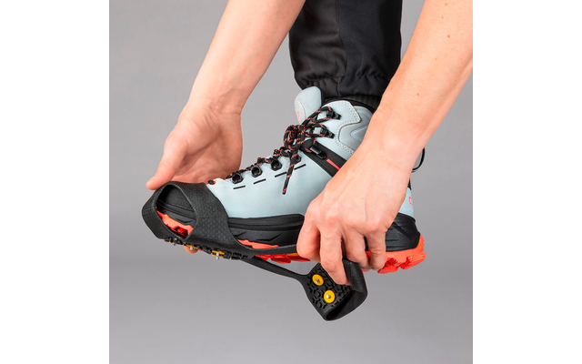 Zwei Hände ziehen ein schwarzes Gummitraktionsgerät mit orangefarbenen Stollen über einen hellblauen Wanderstiefel mit roter Sohle, vor grauem Hintergrund