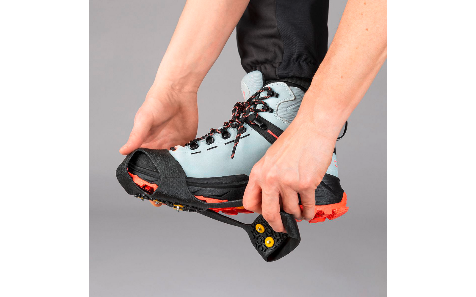 Zwei Hände ziehen ein schwarzes Gummitraktionsgerät mit orangefarbenen Stollen über einen hellblauen Wanderstiefel mit roter Sohle, vor grauem Hintergrund