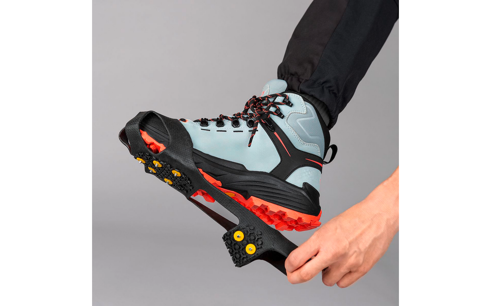 Hand befestigt schwarze Eiskrallen mit gelben Spikes an hellblauem Wanderschuh mit roter Sohle vor grauem Hintergrund