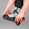 Zwei Hände ziehen ein schwarzes Gummitraktionsgerät mit orangefarbenen Stollen über einen hellblauen Wanderstiefel mit roter Sohle, vor grauem Hintergrund