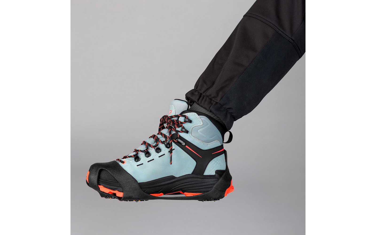 Hellblauer, knöchelhoher Schuh mit schwarzer und orangefarbener Sohle, schwarzem Zehenschutz und schwarz-orange gemusterten Schnürsenkeln, getragen an einem Bein in schwarzer Hose vor hellem grauem Hintergrund