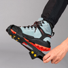 Hand befestigt schwarze Eiskrallen mit gelben Spikes an hellblauem Wanderschuh mit roter Sohle vor grauem Hintergrund