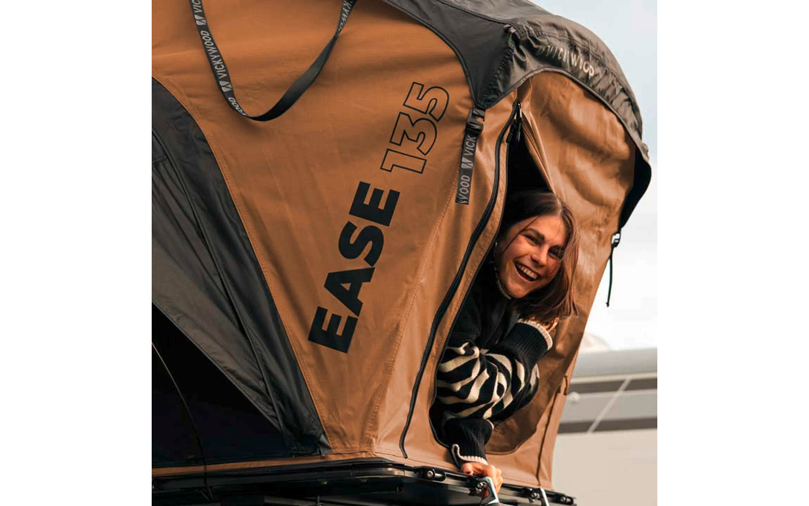 Frau lächelt und schaut aus geöffnetem Dachzelt mit Aufdruck ‚EASE 135‘, braunes Zelt auf Autodach montiert