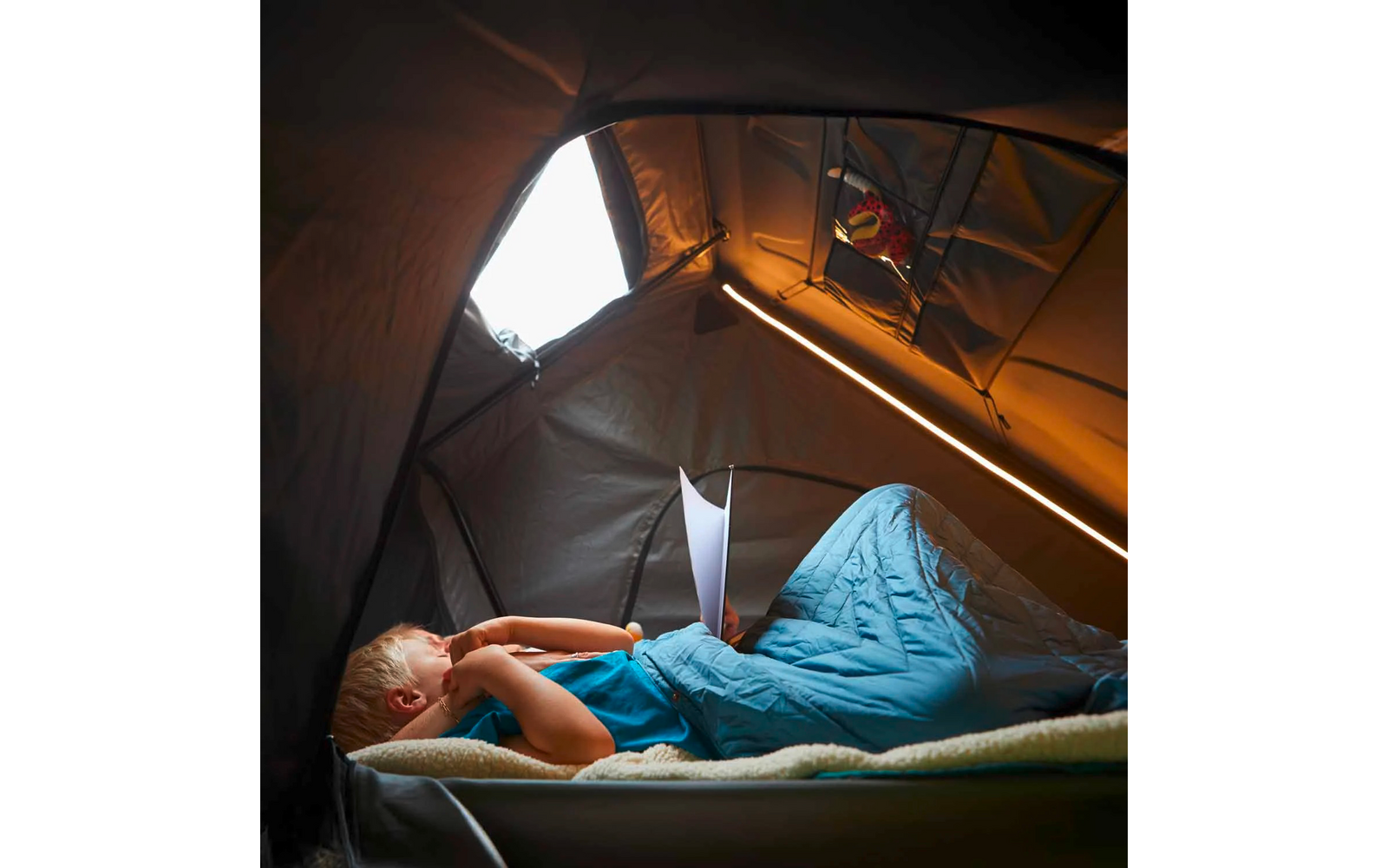 Person liegt in einem blauen Schlafsack auf einer Isomatte im Zelt und hält ein Blatt Papier; LED-Lichtstreifen und geöffnetes Zeltfenster sichtbar