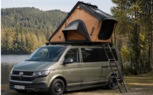 Olive-green Volkswagen camper van parked by a lake, orange rooftop tent open with a ladder, trees and forested hills in the background