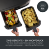 Oben ein schwarzes Gerät, darunter zwei herausnehmbare rechteckige Körbe mit Griffen; linker Korb mit gemischtem Gemüse (Zucchini, Paprika, Champignons), rechter Korb mit gewellten Pommes; zwei Hände halten die Griffe; oben rechts ein dunkler Kreis m
