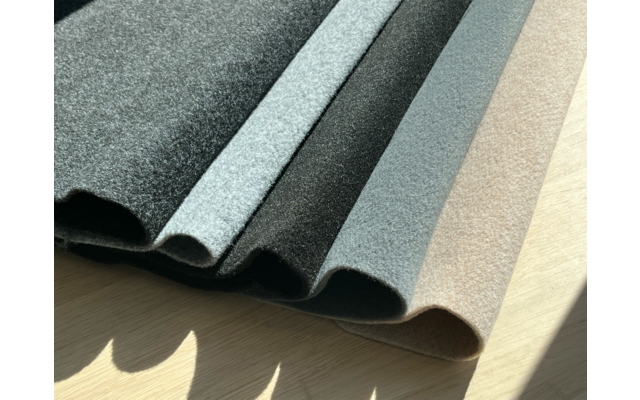 Mehrere wellenförmig gefaltete Stoffbahnen in verschiedenen Grautönen bis Beige auf hellem Holzuntergrund, mit Sonnenlicht und Schatten