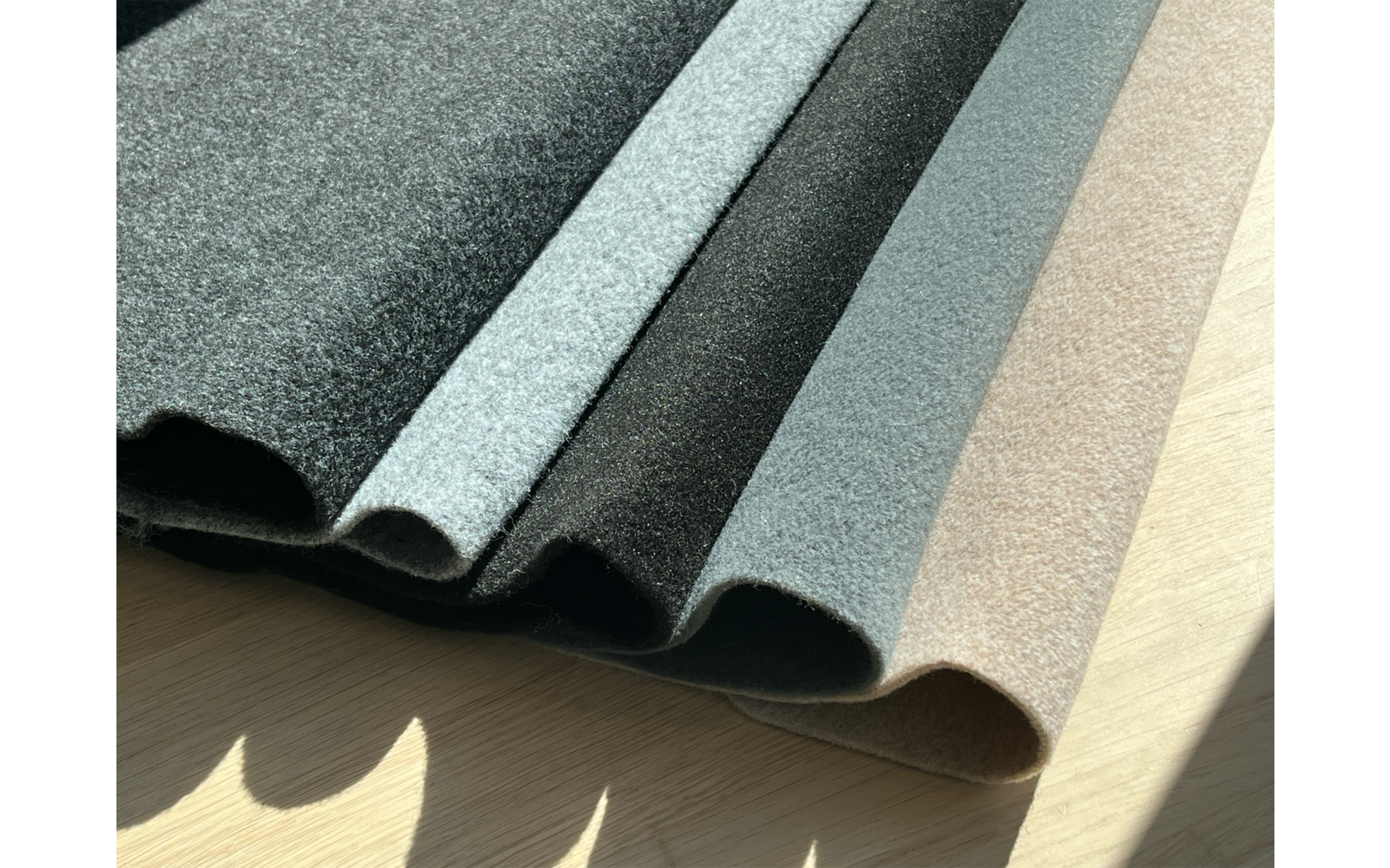 Mehrere wellenförmig gefaltete Stoffbahnen in verschiedenen Grautönen bis Beige auf hellem Holzuntergrund, mit Sonnenlicht und Schatten