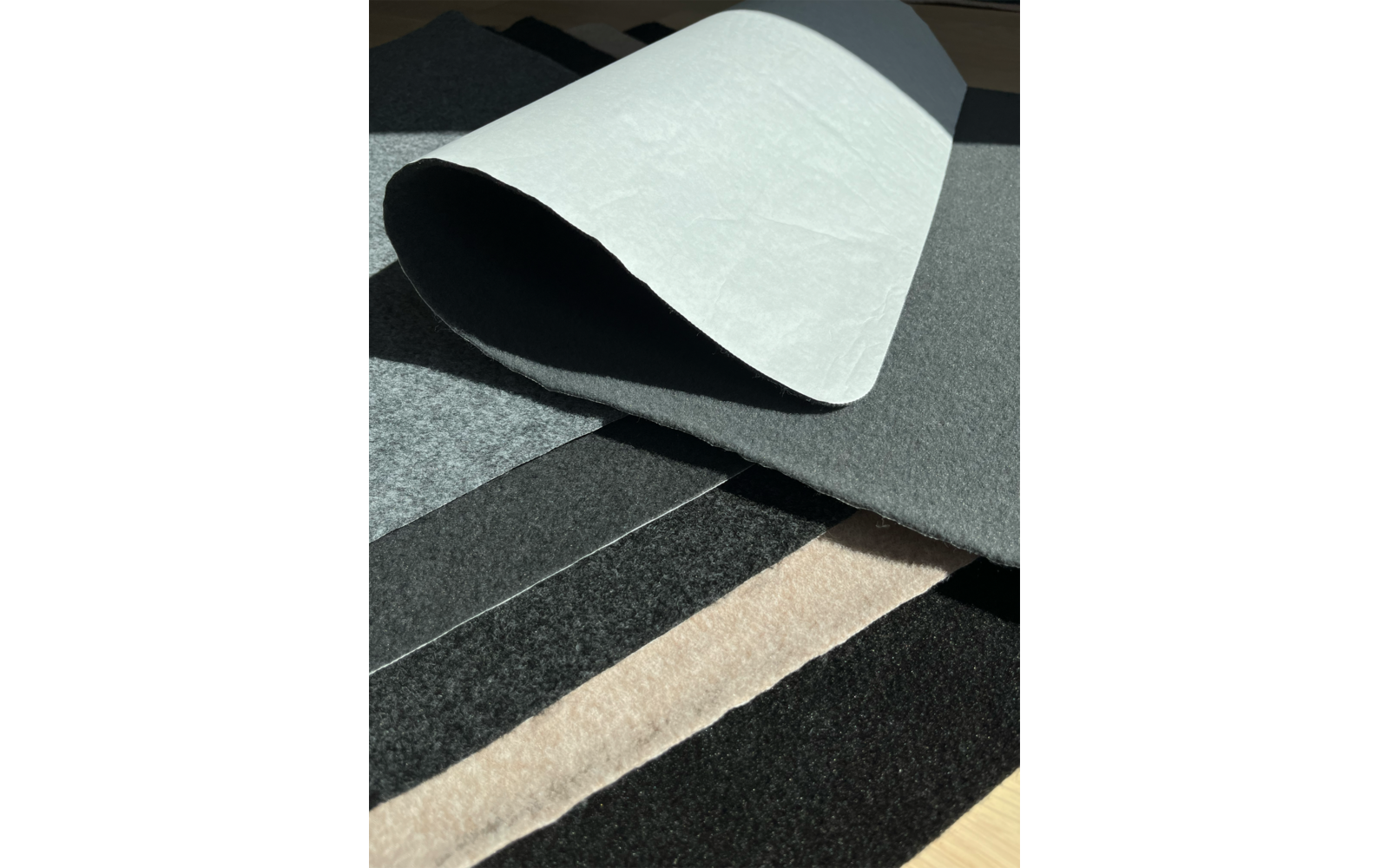 Aufgerollene weiße Bahn liegt über mehreren flachen Lagen in Grau, Schwarz und Beige auf hellem Holz mit Schatten