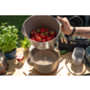 Handen houden een beige kom met aardbeien die in een beige pan worden gegoten; op de houten tafel staan een basilicumplant, een potje met rood-wit geruit deksel, een rasp en andere keukenspullen