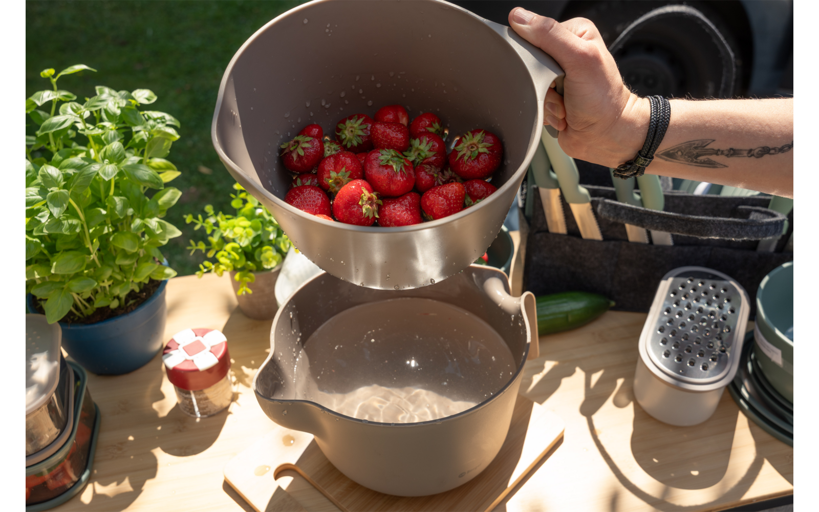 Handen houden een beige kom met aardbeien die in een beige pan worden gegoten; op de houten tafel staan een basilicumplant, een potje met rood-wit geruit deksel, een rasp en andere keukenspullen