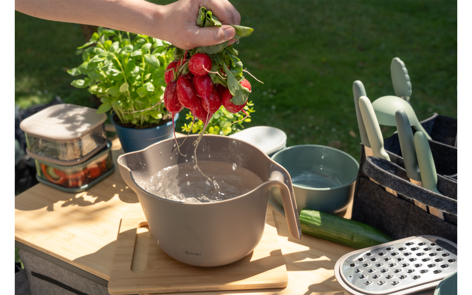 Hand houdt een bosje rode peulen boven een kom, er druppelt water van de peulen; op de houten tafel staan kruiden in pot, een komkommer, een rasp en keukengerei