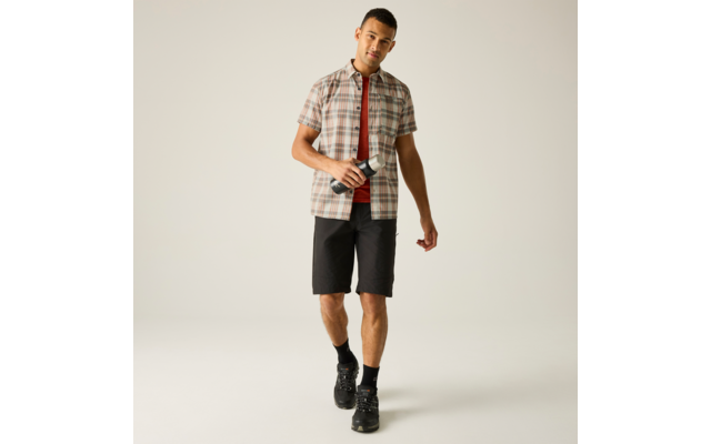 Mann trägt kariertes Kurzarmhemd über rotem T‑Shirt, schwarze Shorts, schwarze Socken und Schuhe, geht nach vorn und hält in der rechten Hand ein kleines schwarzes Gerät