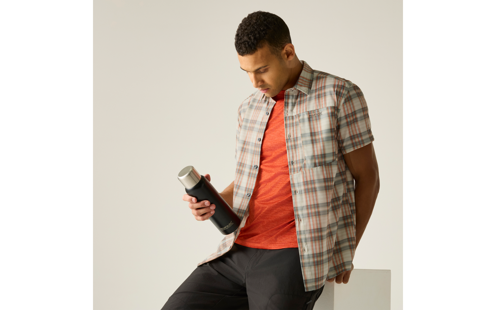 Mann in kariertem Hemd und rotem T‑Shirt sitzt, hält eine schwarze Trinkflasche und schaut auf die Flasche