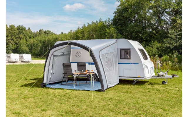 Caravane blanche avec auvent gris; dans l'auvent une table et deux chaises; pelouse, arbres et ciel bleu avec des nuages