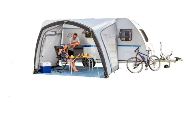 Caravane avec auvent sur l'herbe près de la plage, deux personnes assises à une table, glacière, deux vélos, plage de sable et rochers en arrière-plan, ciel bleu