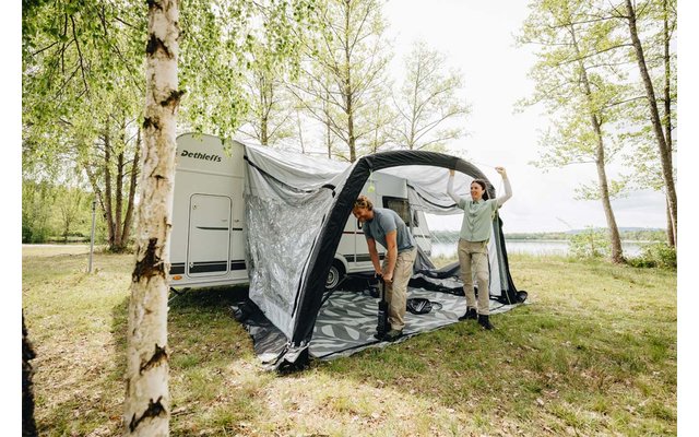 Dos personas colocan un toldo hinchable en una caravana blanca sobre césped junto a abedules y un lago